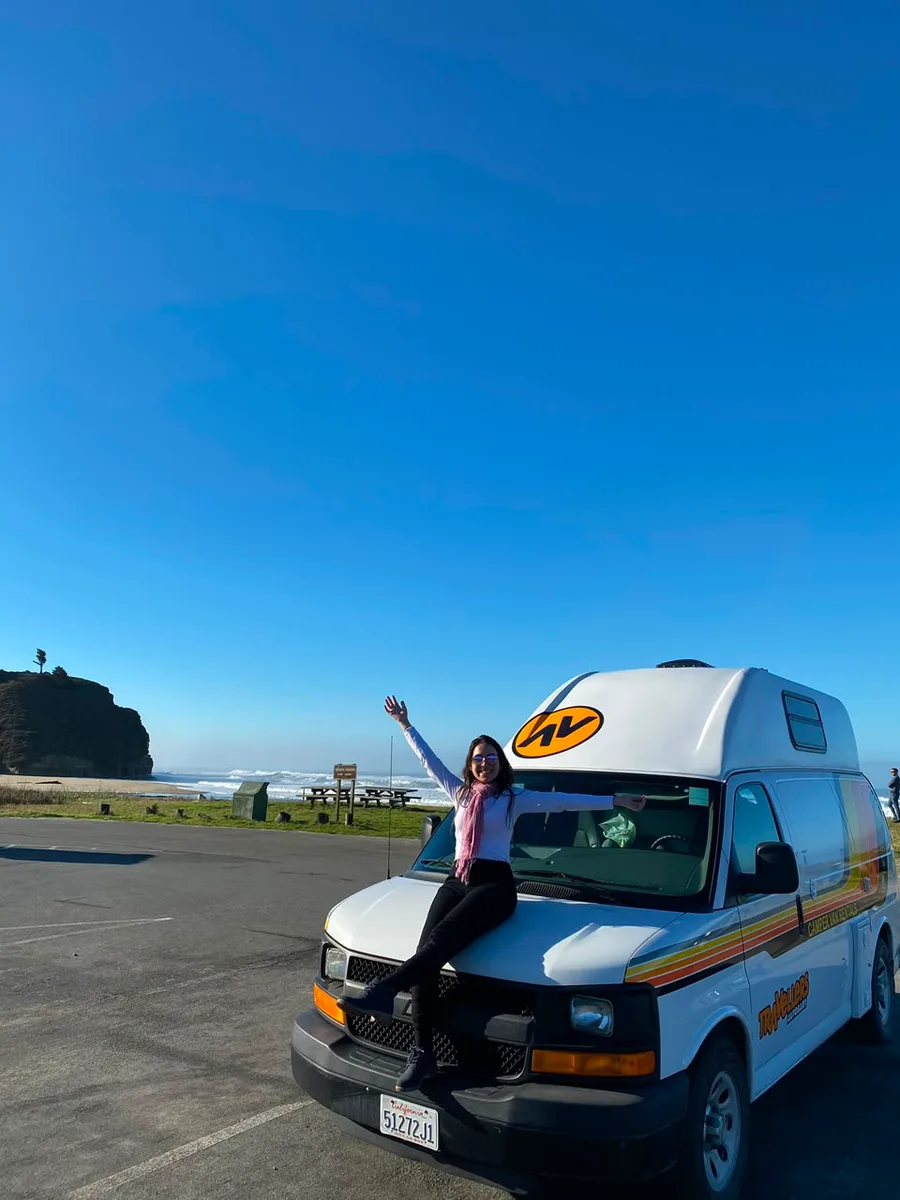 Julia sentada na frente de uma van de viagem, com o braço erguido, à beira da estrada