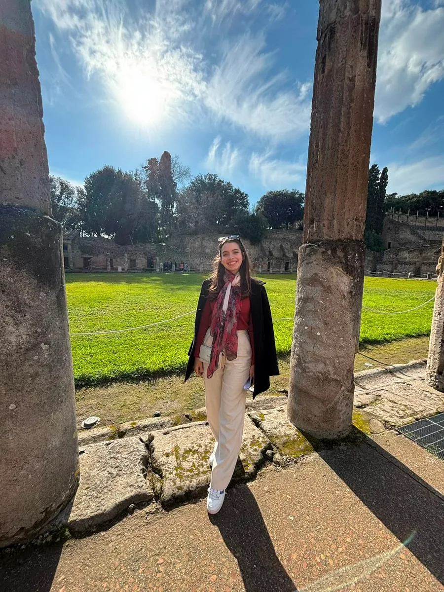 Julia em pé entre colunas antigas de um sítio arqueológico, num dia de sol
